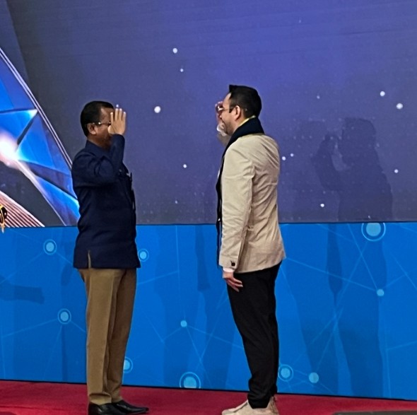 Two men stand on a stage facing each other; one raises his hand in a greeting or gesture while the other listens. Both wear smart casual attire against a blue, tech-themed backdrop.